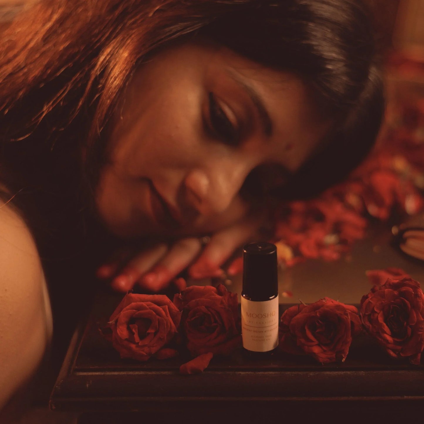 Woman lying on a bed of red roses with a perfume oil bottle of perfume next to her.