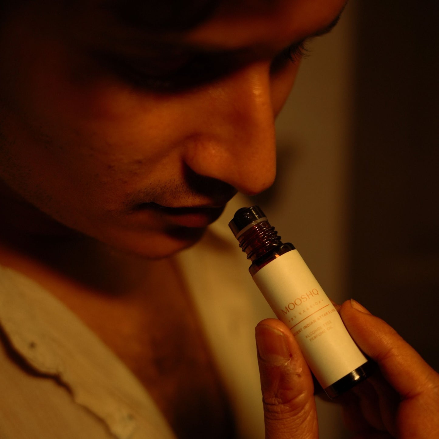 A man holding a bottle of Mooshq perfume oil close to their nose his a warm, orange glow.
