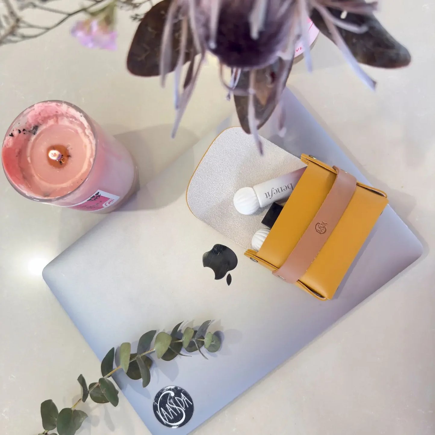 Laptop with a yellow and pink brown perfume oil pouch on a white surface with a pink candle and plant.