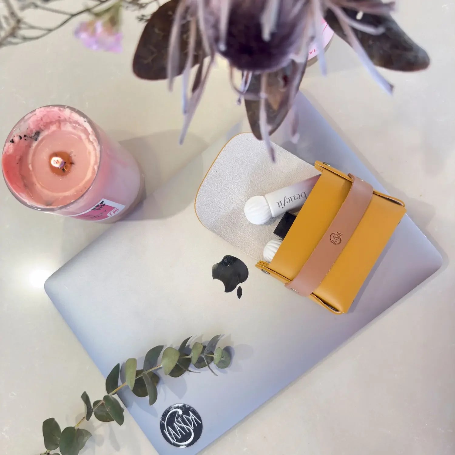 Laptop with a yellow and pink brown perfume oil pouch on a white surface with a pink candle and plant.
