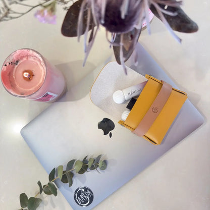 Laptop with a yellow and pink brown perfume oil pouch on a white surface with a pink candle and plant.