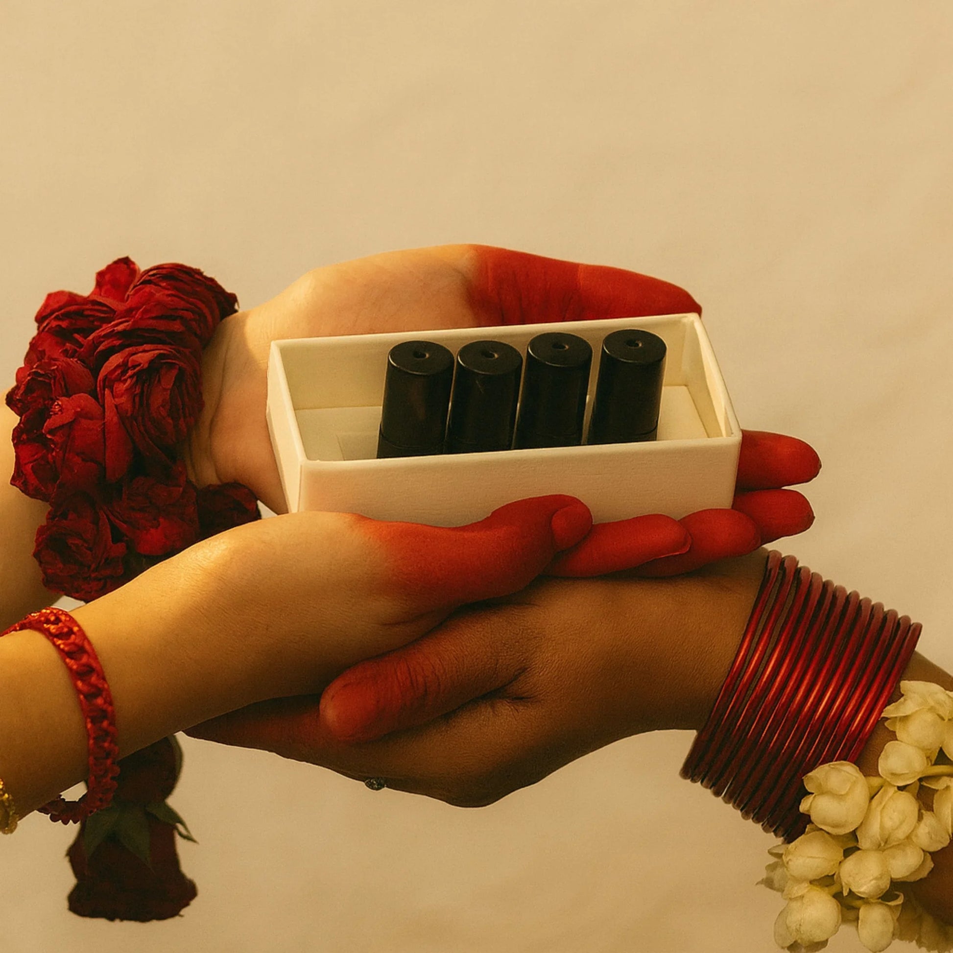 Hands holding a white box with perfume oil bottles, surrounded by red and white flowers on a beige background.