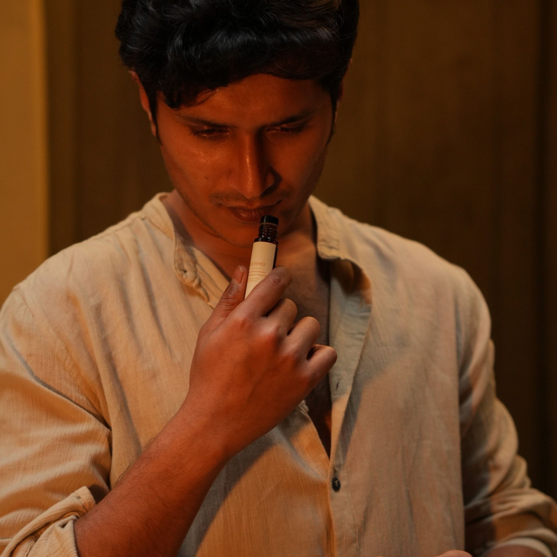 Man in a beige shirt holding Mooshq Perfume oil close to his nose against a brown background