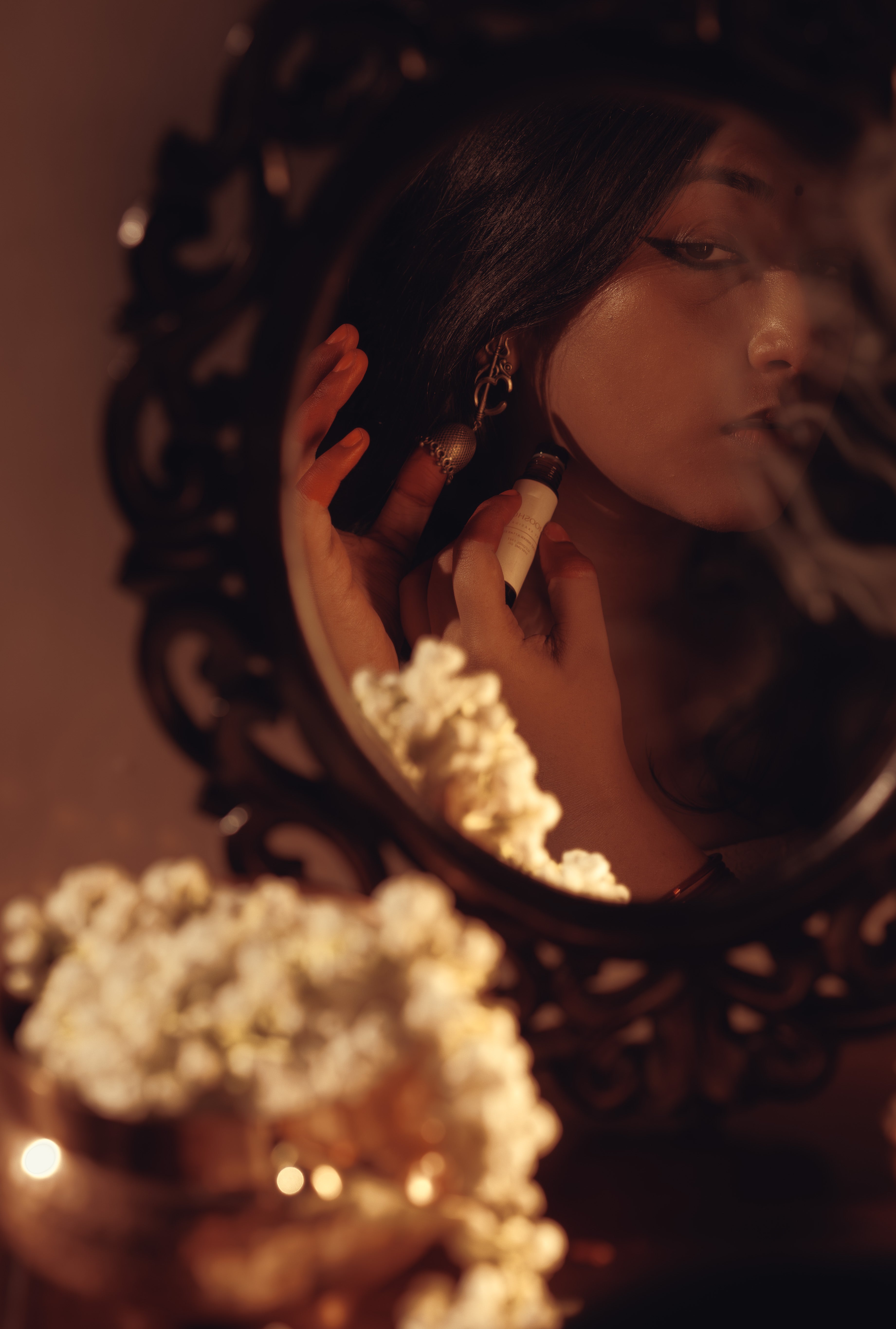 Close-up of a woman applying perfume oil  against a decorative mirror reflecting their face, surrounded by soft lighting and jasmins.