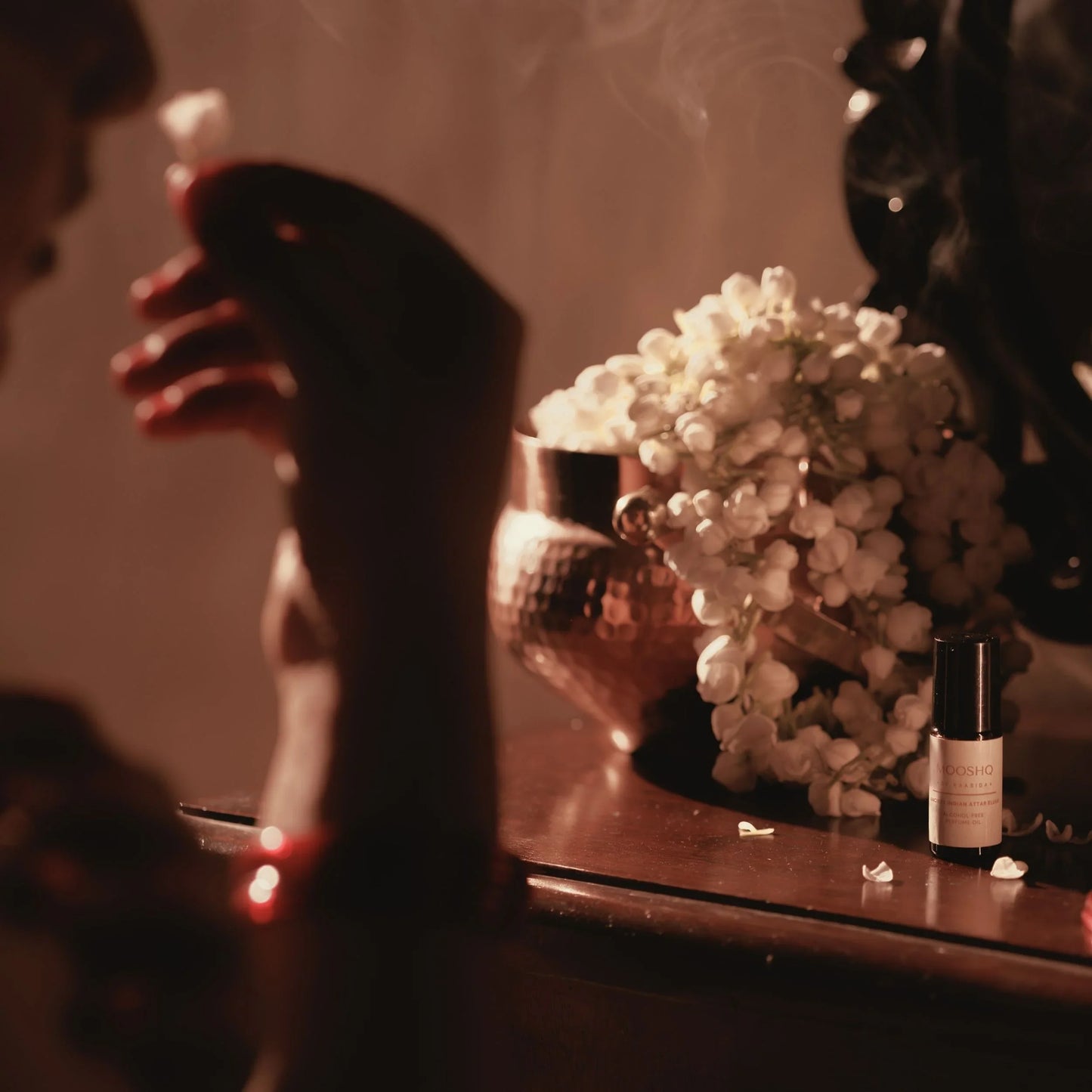 Perfume Oil bottle on a reflective surface with jasmin flowers and a hand in the foreground