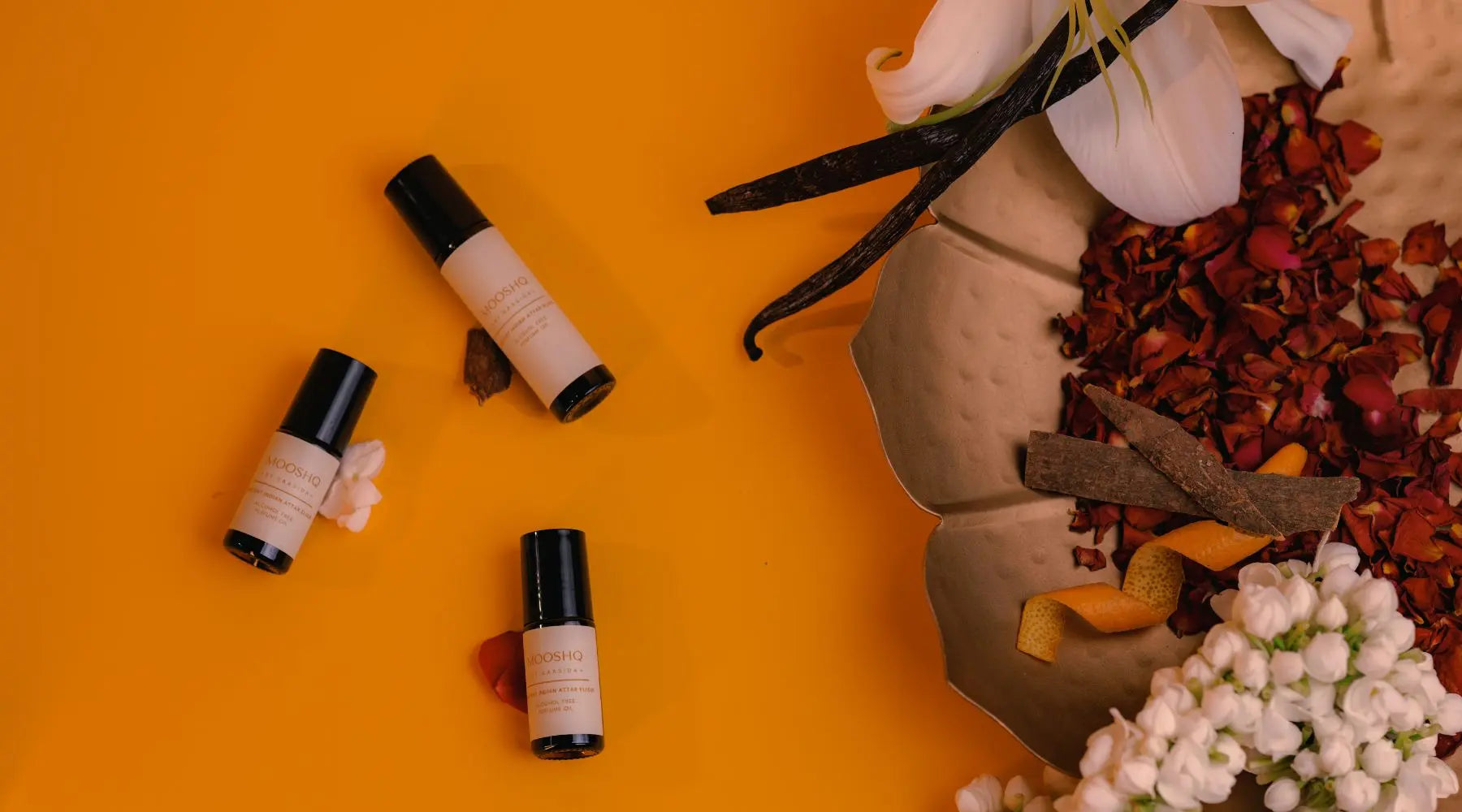 Vanilla beans and rose petals and jasmin flowers on an orange background along with 3 bottles of Mooshq Perfume Oil Attar
