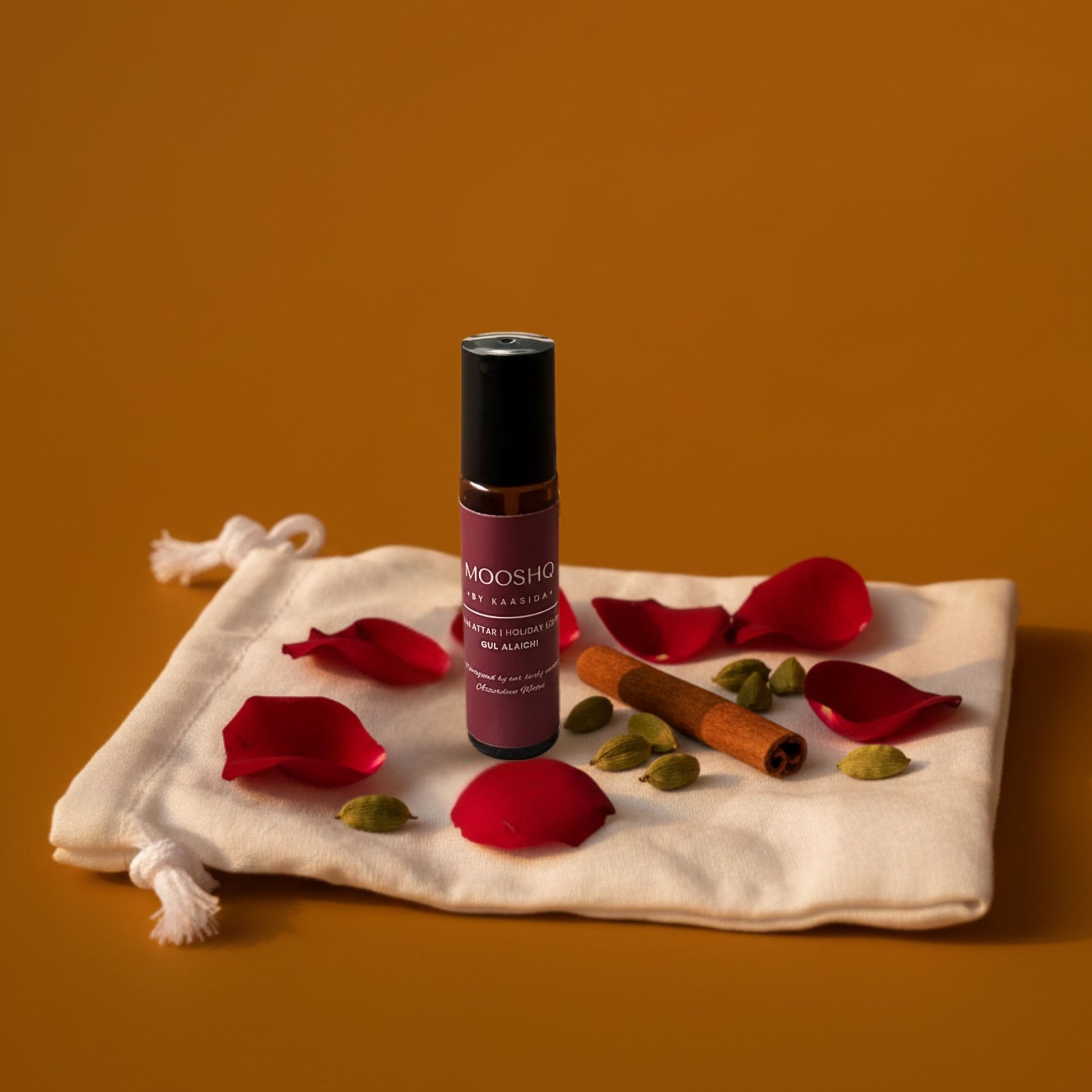 Mooshq Perfume Oil with red rose petals and cinnamon sticks on a fabric pouch against an orange background