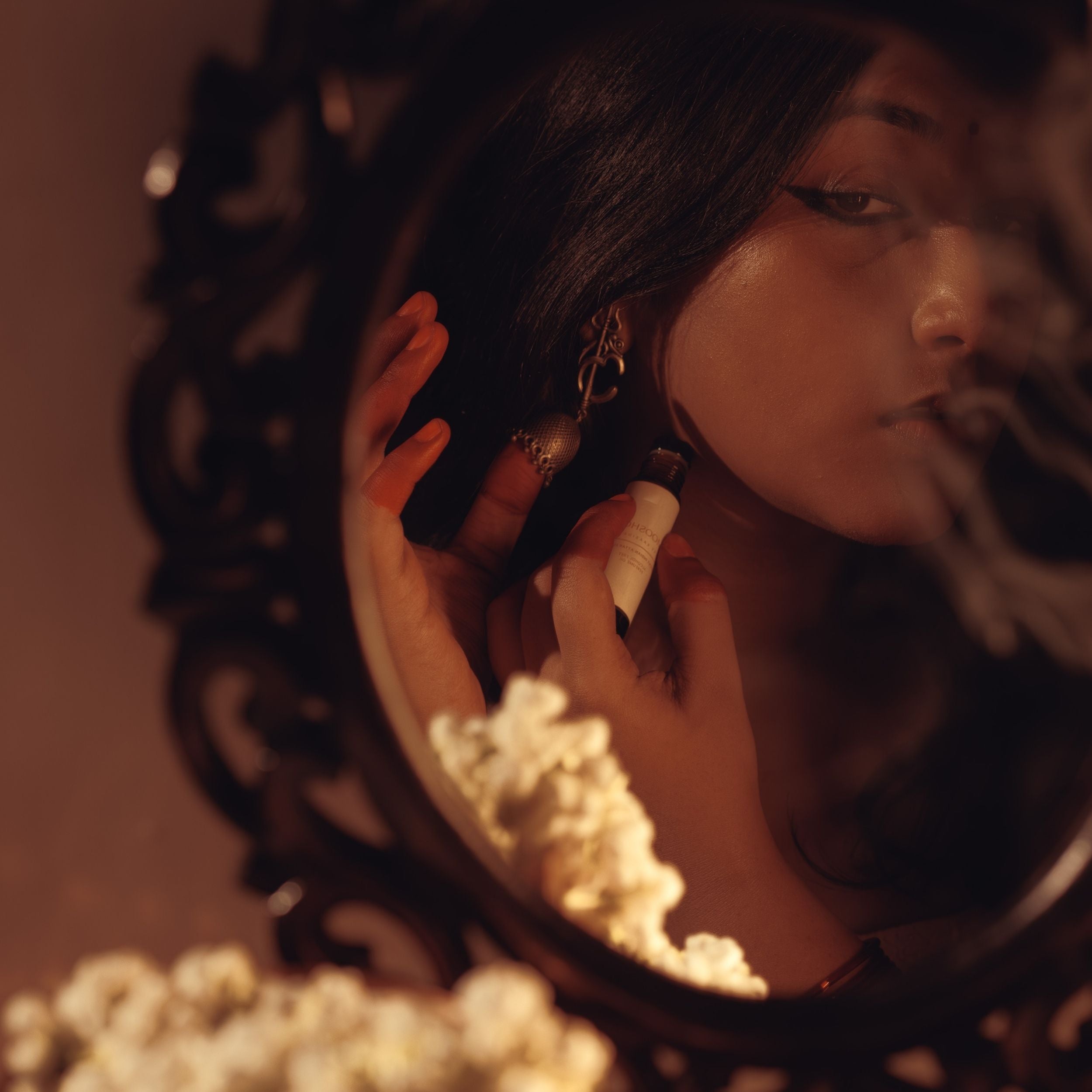 Woman applying perfume oil behind her ear before a decorative mirror and jasmin flowers in the foreground.