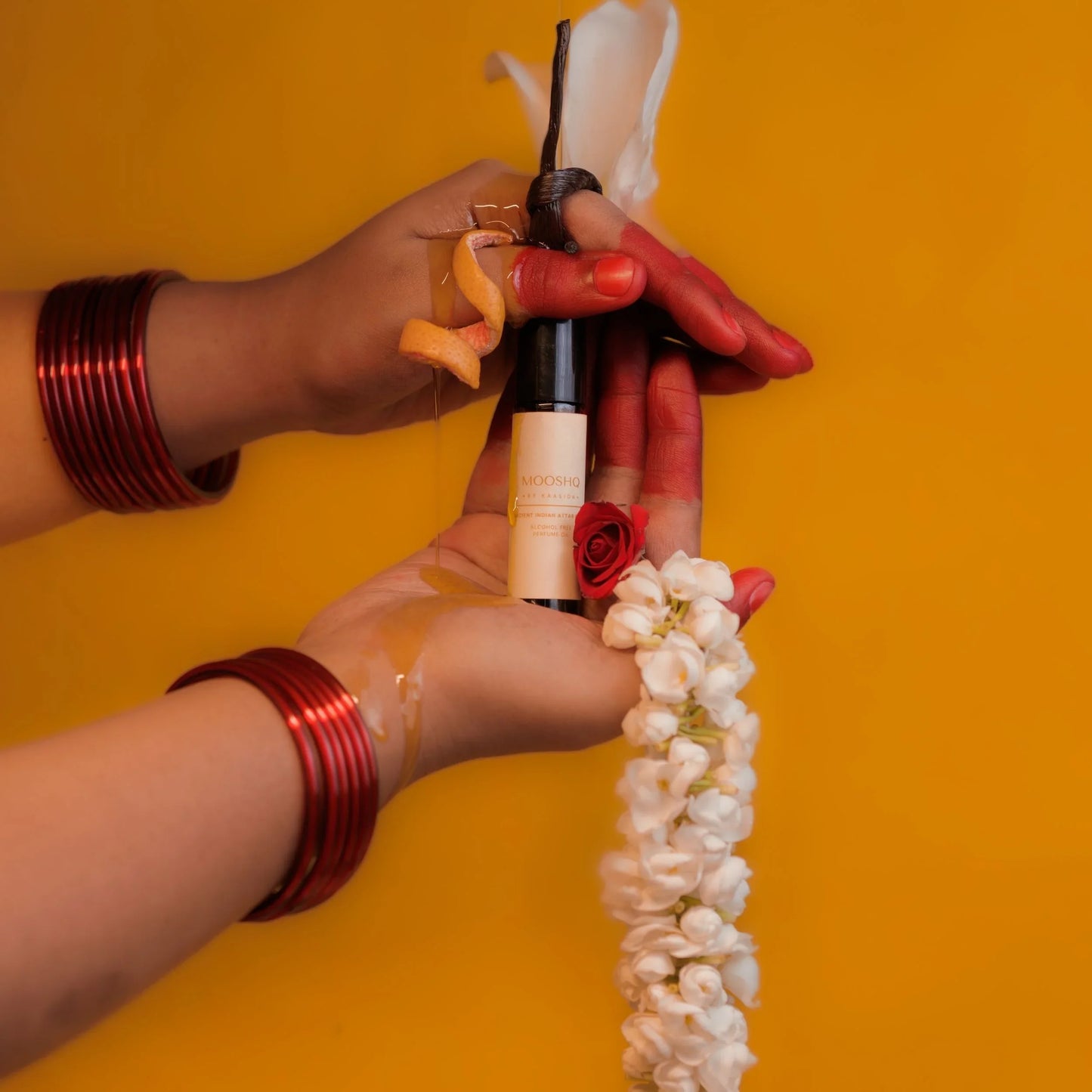 Hands with red bangles holding a perfume oil bottle and a flower garland against an orange background