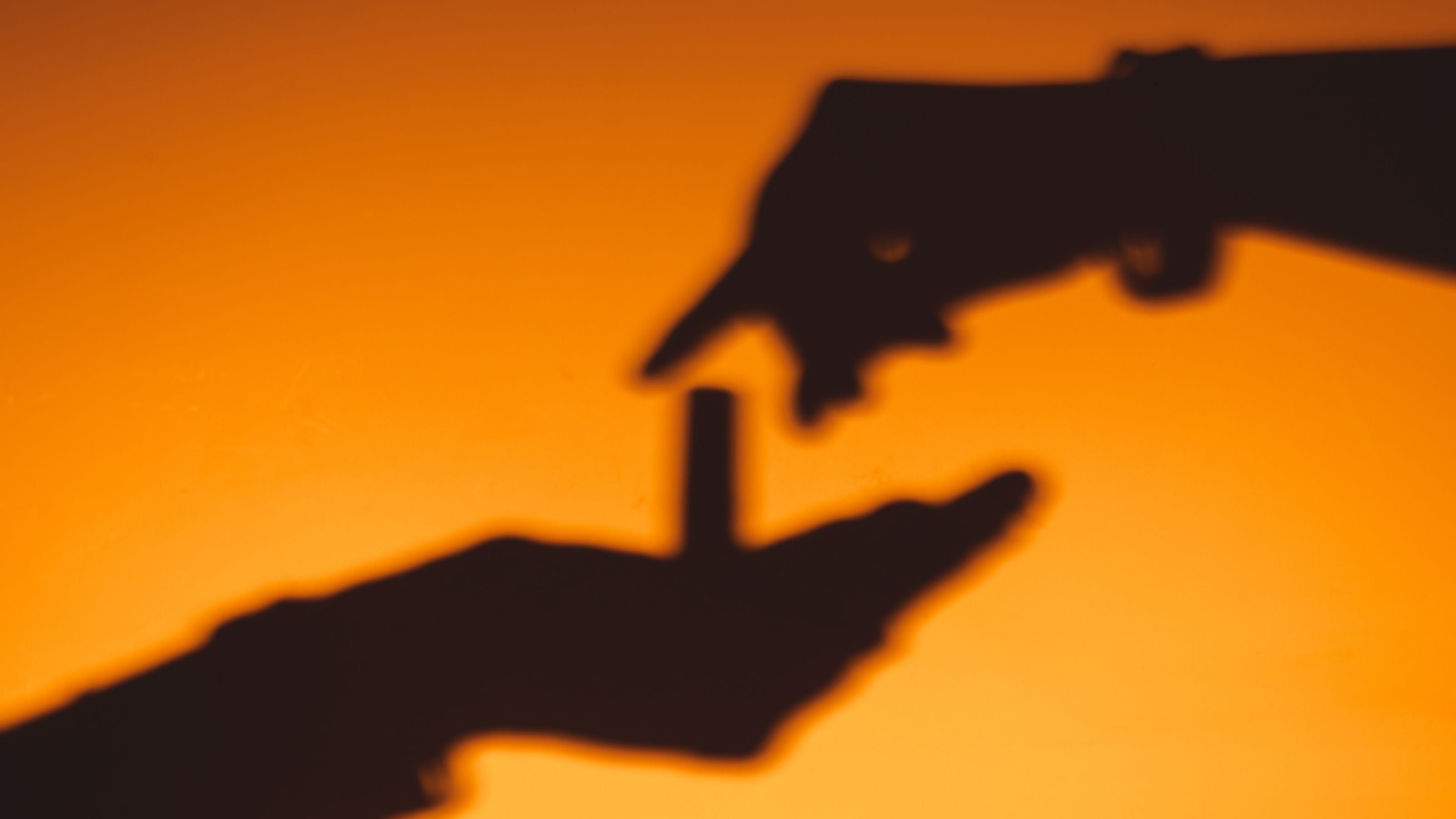 Shadow of a hand holding a perfume oil bottle with another hand reaching out against an orange background