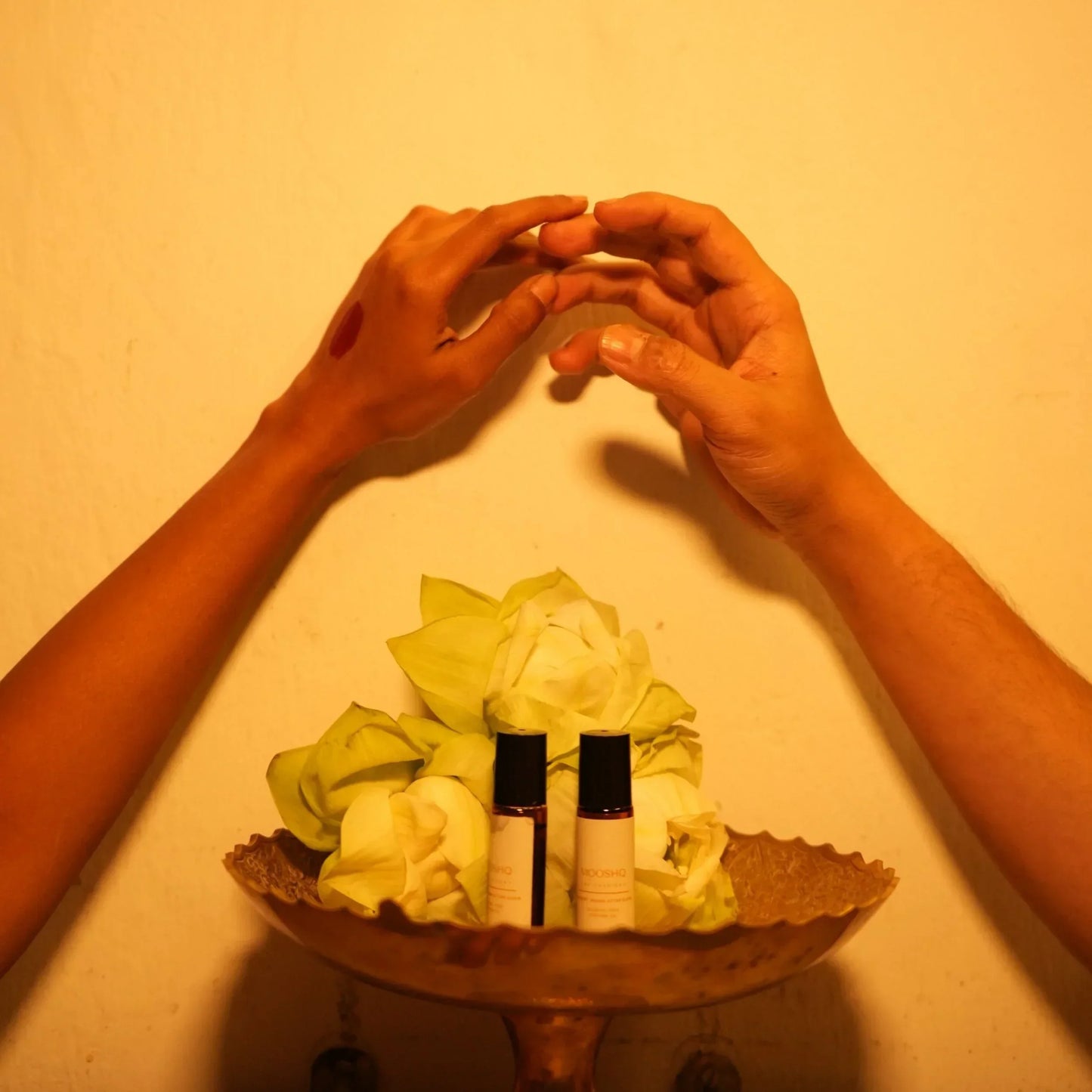 Two hands forming a heart shape above Mooshq perfume oil bottles with lotus in the center on a beige background.