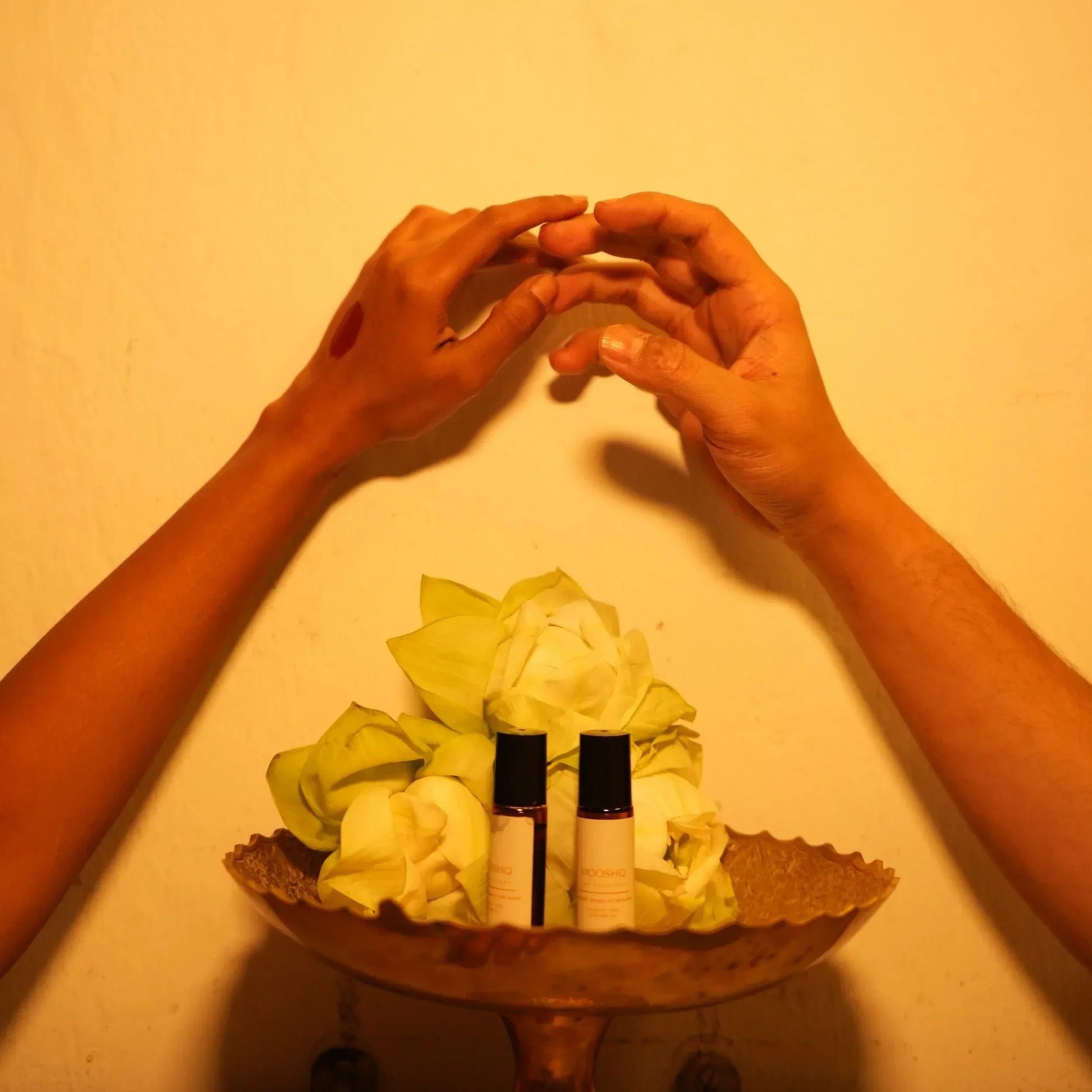 Two hands forming a heart shape above Mooshq perfume oil bottles with lotus in the center on a beige background.