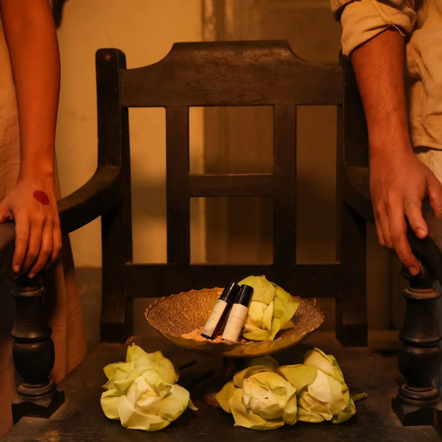 Two hands on a wooden chair with a bowl of flowers and Mooshq Perfume Oil bottles
