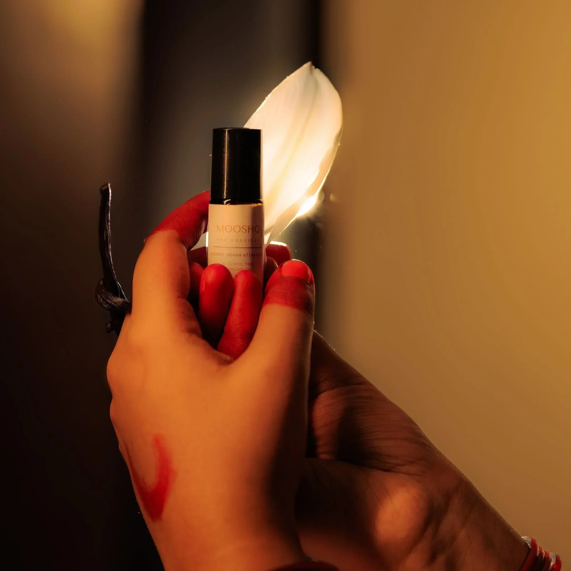 Hand holding a perfume oil with lily petal and vanilla strand with a dark background
